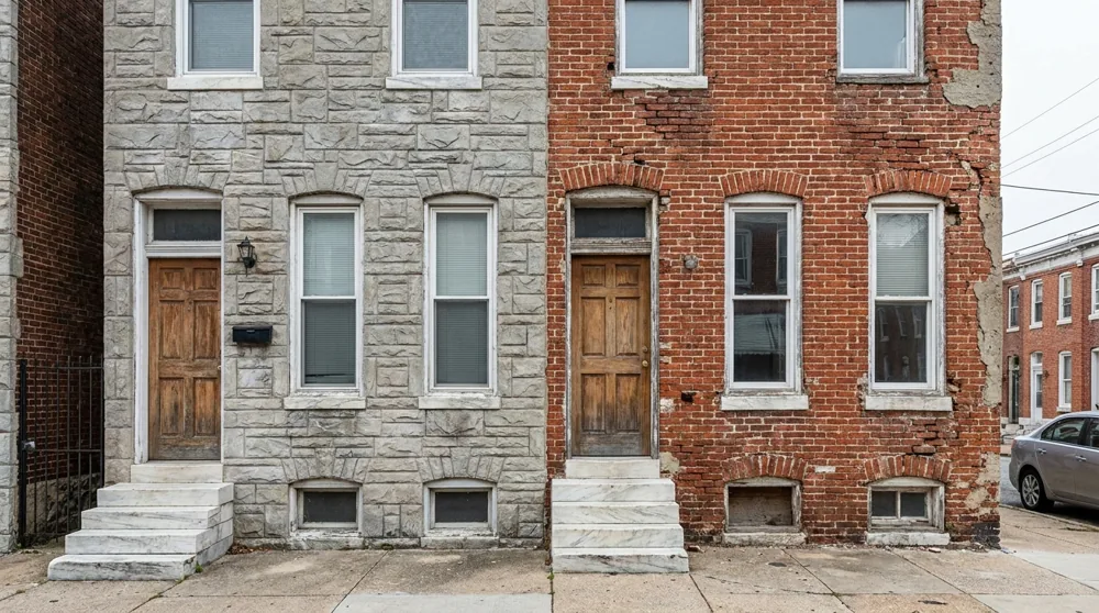 Two adjacent Baltimore rowhomes, one still wearing grey formstone and the neighbor with formstone removed showing exposed weathered red brick