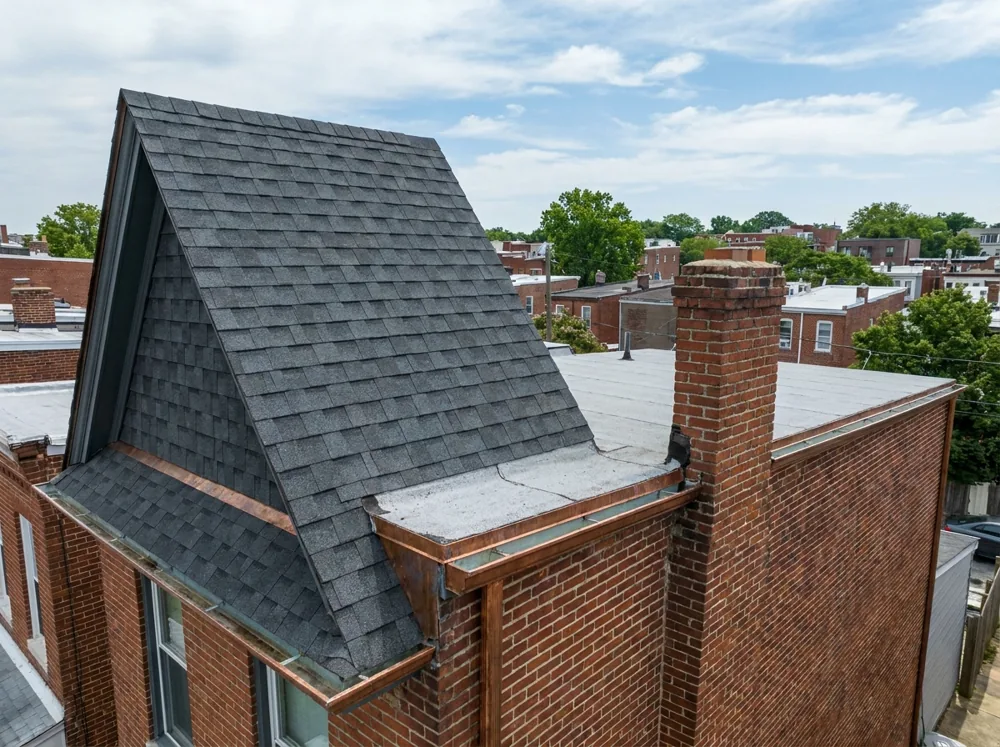 A Baltimore home with a sloped front section of dark gray architectural asphalt shingles transitioning to a flat low-slope rear roof, with a brick chimney and gutters